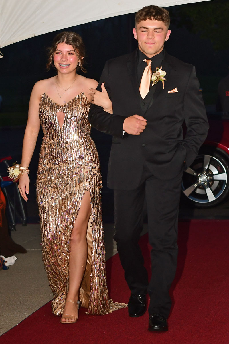 Two teenagers in formal attire walking arm-in-arm on a red carpet at prom: girl in a gold sequined gown, boy in a black suit with a peach tie and boutonniere.