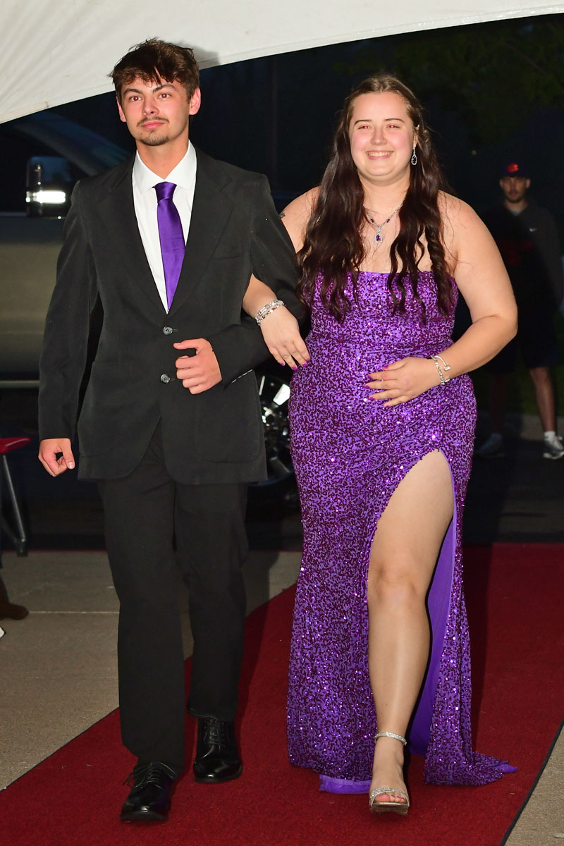 Couple walking arm-in-arm on a red carpet; woman in a purple sequin gown with a high slit, man in a black suit with a purple tie.