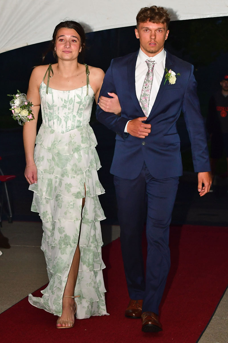 Couple walking arm-in-arm on a red carpet; woman in a tiered green floral gown, man in a navy suit with boutonniere.