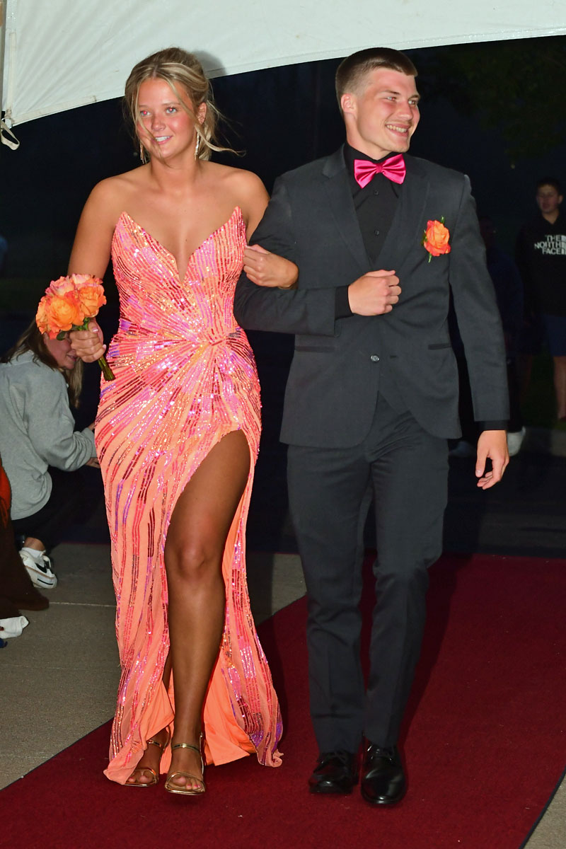 Couple walking a red carpet: girl in a pink sequin gown with a high slit holding orange bouquet, man in black suit with pink bow tie and boutonniere.