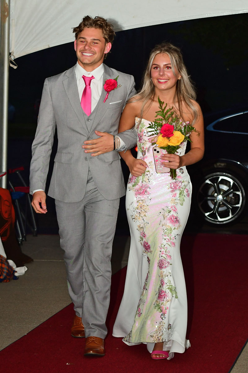 Smiling couple walking arm-in-arm on a red carpet: man in a gray suit with pink tie and boutonniere, woman in a floral gown holding a bouquet of roses.