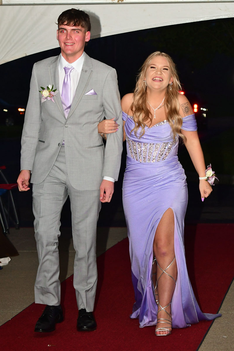 A smiling couple walks arm-in-arm on a red carpet; man wears a light gray suit with lavender tie, boutonniere, and black shoes, woman wears a lavender off-shoulder gown with a high slit and silver accessories behind him.
