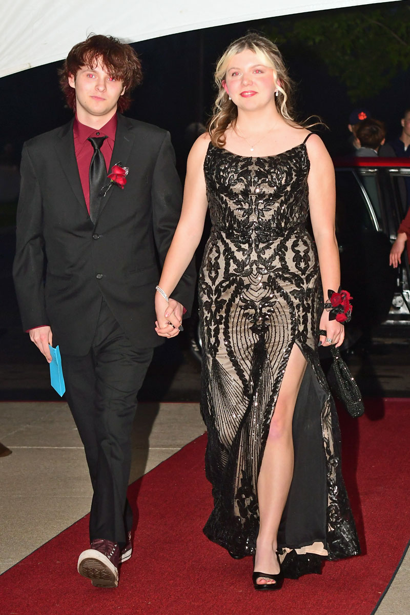Couple walking hand in hand on a red carpet at a formal event; man in black suit with burgundy shirt and boutonniere, woman in black lace gown with a high slit and clutch.",