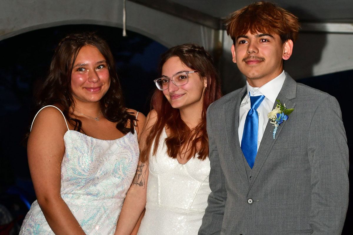 Three friends in formal attire posing together: two girls in white dresses and a boy in a gray suit with a blue tie, smiling at the camera.