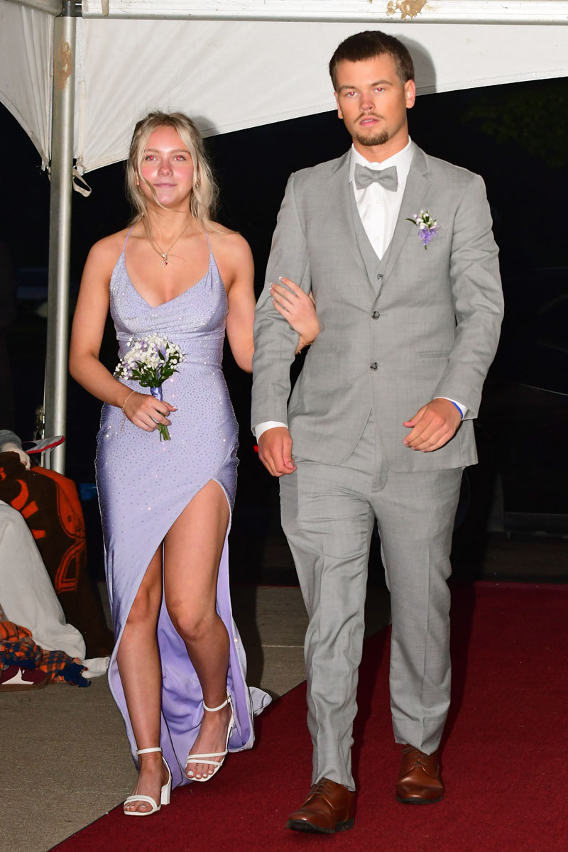 Young couple walking arm in arm on a red carpet; woman in a lavender gown holding a small bouquet, man in a light gray suit and bow tie.