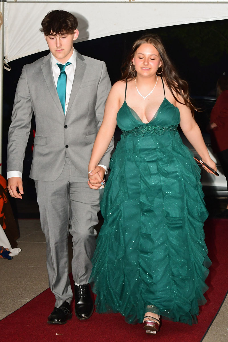 Young man in a gray suit with teal tie and a young woman in an emerald-green gown walk hand in hand on a red carpet.