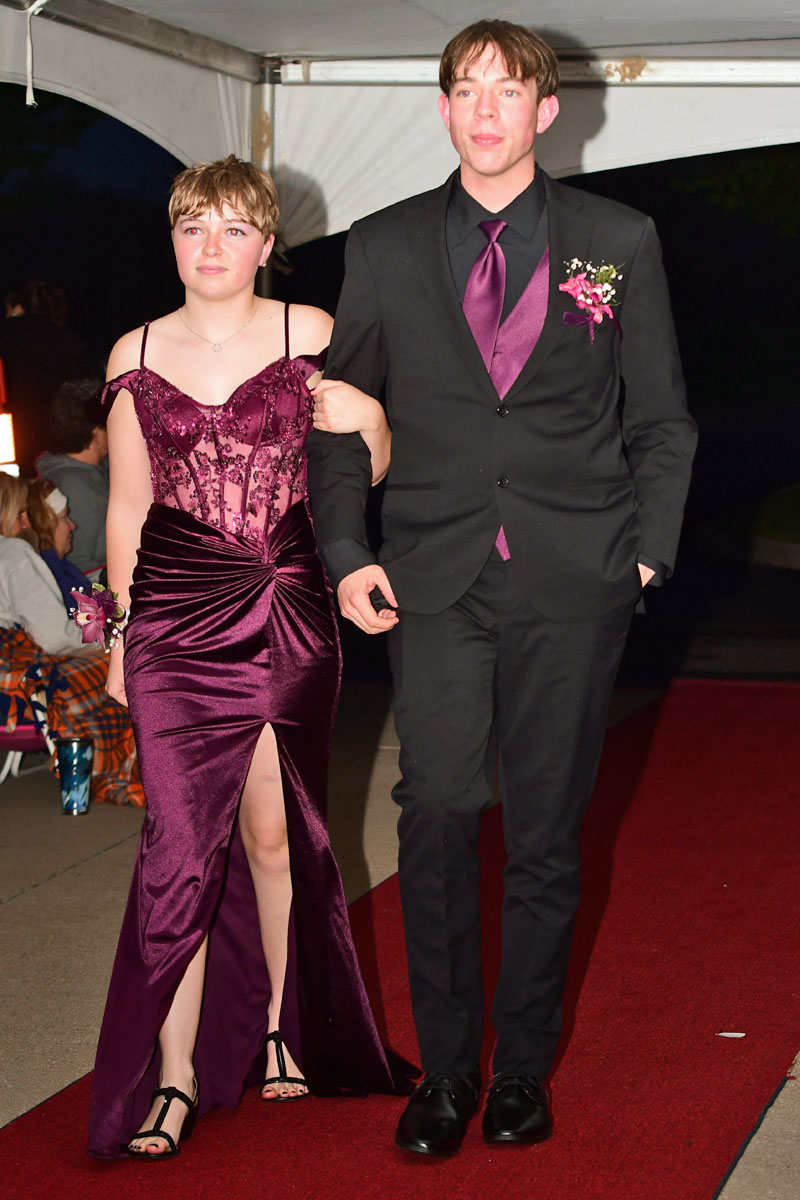 Two teens walk arm-in-arm on a red carpet; girl in a burgundy lace gown, guy in a black suit with burgundy tie and corsage.