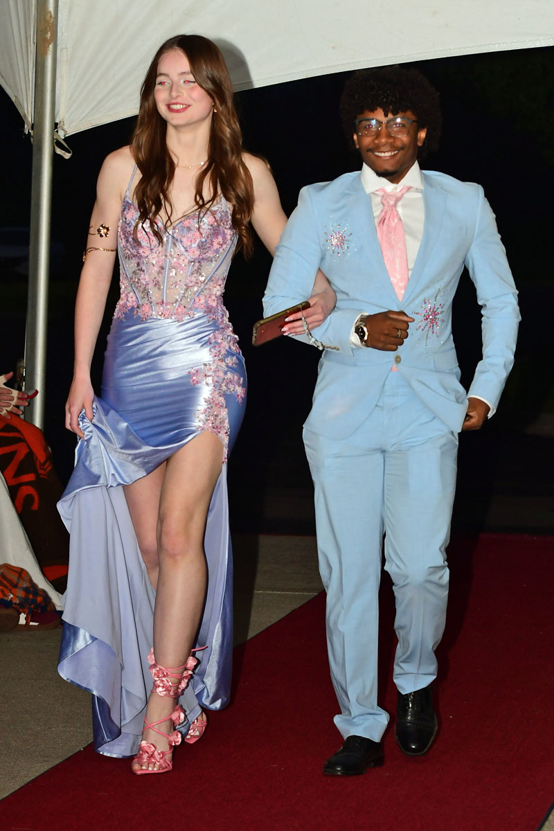 Two young adults walk arm-in-arm on a red carpet; woman in a lilac gown with pink embellishments and pink heels, man in a light blue suit with a pink tie smiles at the camera.