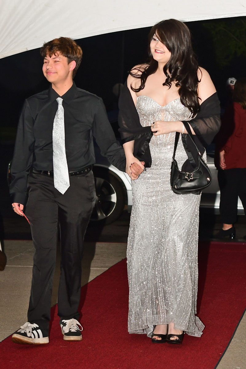 Young couple walking hand in hand on a red carpet; girl in a silver beaded gown with black shawl and purse, boy in black shirt and light tie, both smiling.