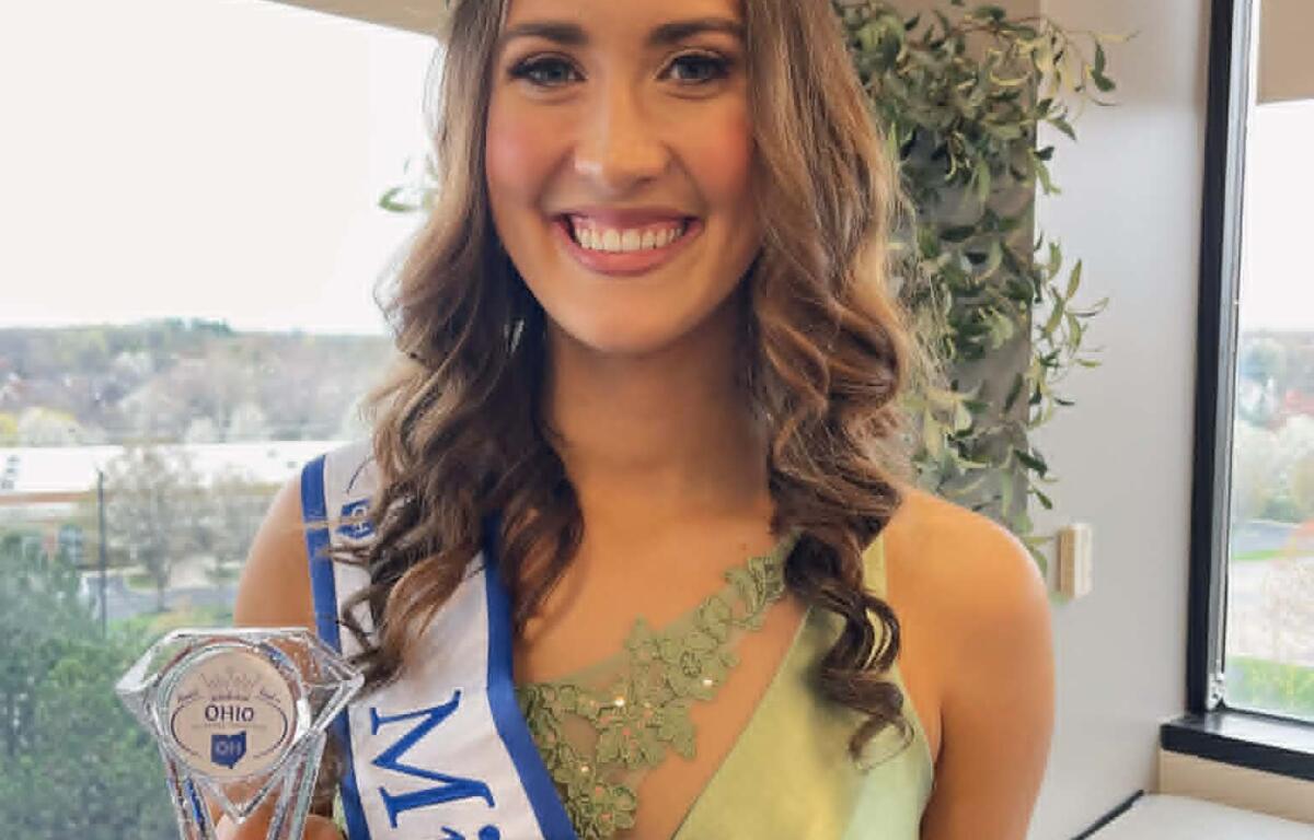 Smiling pageant winner wearing a crystal crown and sash, holding a trophy in front of a window.