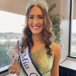 Smiling pageant winner wearing a crystal crown and sash, holding a trophy in front of a window.