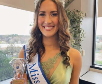 Smiling pageant winner wearing a crystal crown and sash, holding a trophy in front of a window.