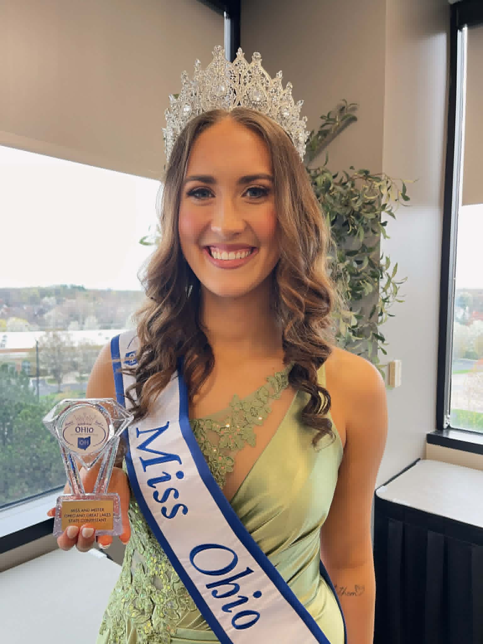 Smiling pageant winner wearing a crystal crown and sash, holding a trophy in front of a window.