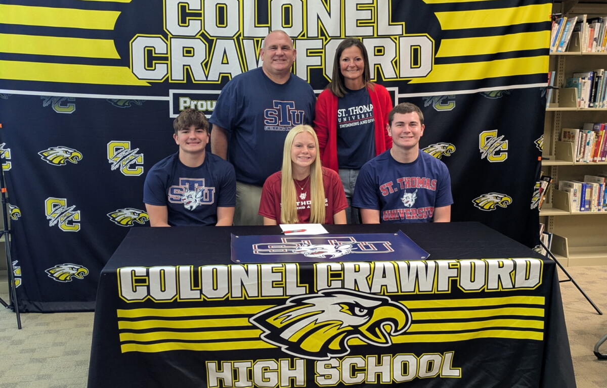 Group of five people posing for a photo in front of a Colonel Crawford High School banner, wearing STU and university-themed shirts in a library setting.
