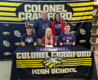 Group of five people posing for a photo in front of a Colonel Crawford High School banner, wearing STU and university-themed shirts in a library setting.