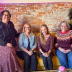 Five women sit on a yellow couch against an exposed brick wall, smiling at the camera in a casual indoor space.