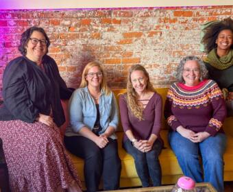 Five women sit on a yellow couch against an exposed brick wall, smiling at the camera in a casual indoor space.