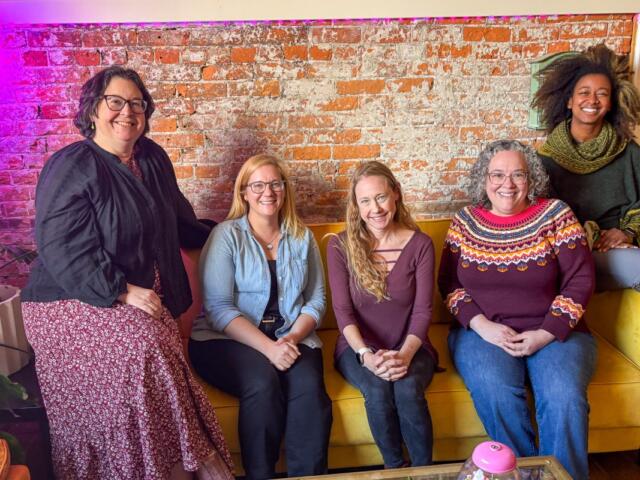 Five women sit on a yellow couch against an exposed brick wall, smiling at the camera in a casual indoor space.