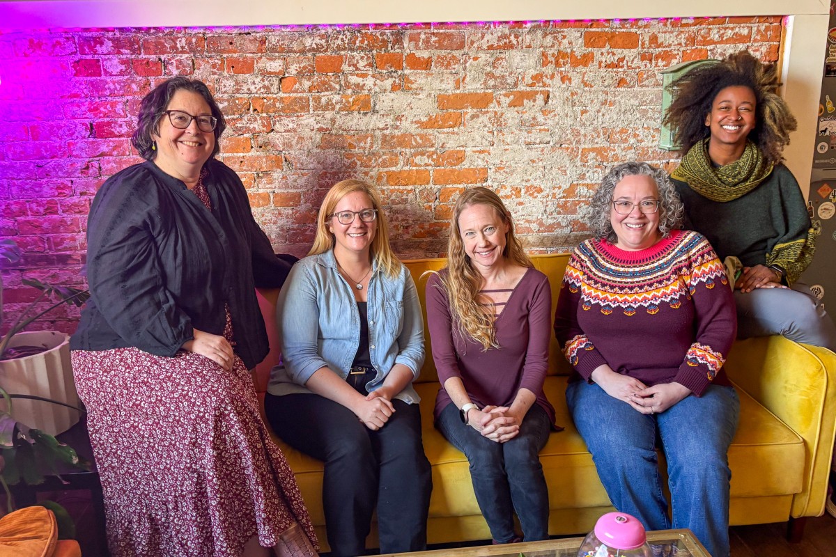 Five women sit on a yellow couch against an exposed brick wall, smiling at the camera in a casual indoor space.