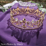 Gold ornate crown with jewels rests on purple fabric, casting a decorative shadow.