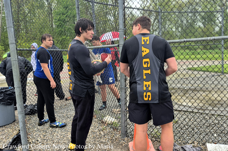 Two young men in black Eagles jerseys stand by a chain-link fence, with one holding a softball and talking to a teammate.