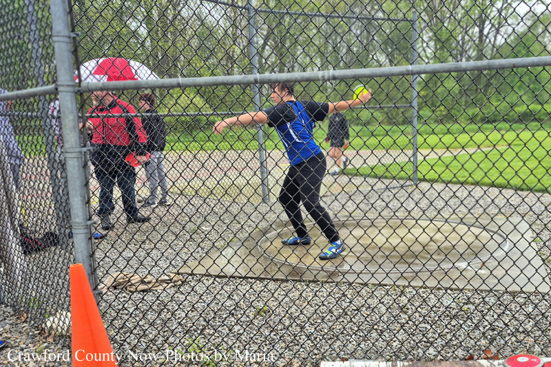 Athlete in a blue shirt throws a shot put inside a fenced throwing circle at a park, with spectators nearby.