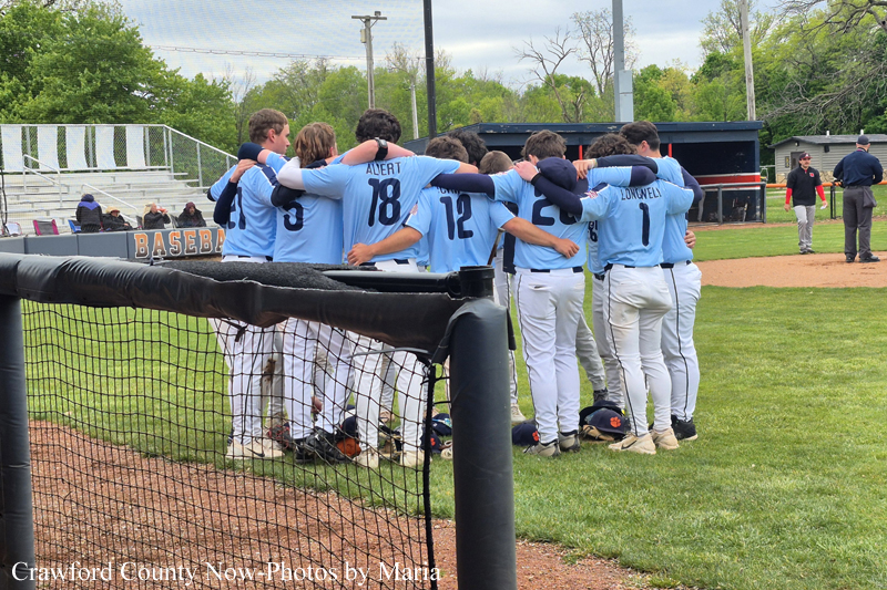 Youth baseball team in light blue jerseys forming a huddle on the field near the dugout.
