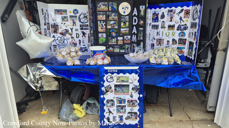 Display booth in a garage with a blue tablecloth, baked snacks, and three large photo-collage boards celebrating a student named Austin.