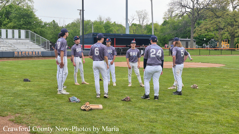 Youth baseball team in purple uniforms forming a huddle on a green field; gloves and equipment lie on the grass, with empty bleachers and a 'BASEBALL' sign in the background.