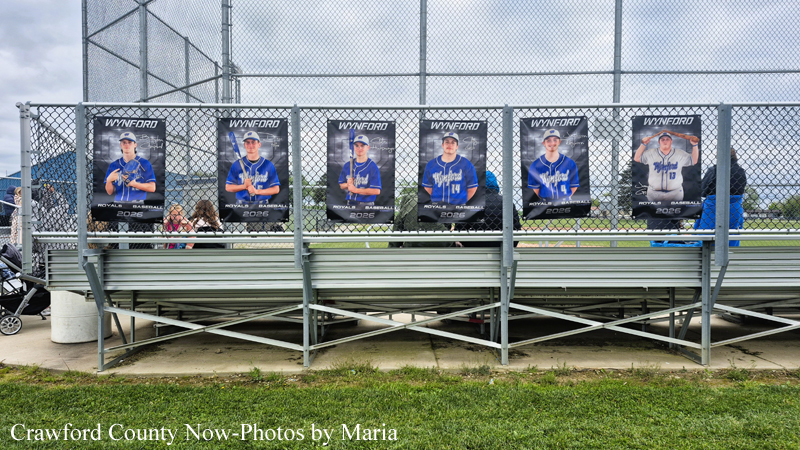 Six baseball posters attached to bleachers behind a chain-link fence at a ballpark, with spectators in the background; caption reads Crawford County Now—Photos by Maria.