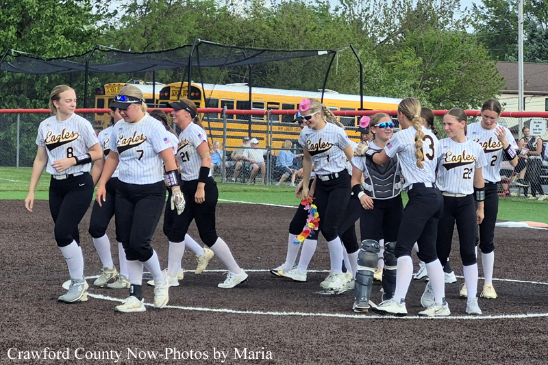 Softball team walking together on the infield after a game, wearing white pinstriped jerseys that say Eagles and black pants; school buses and a fence visible in the background.