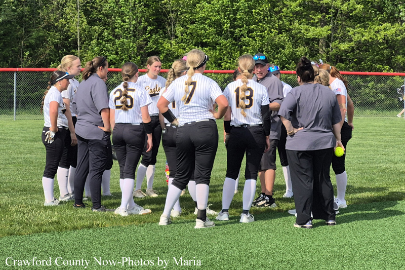 Softball team huddle on a grassy field, players in white pinstriped jerseys with numbers, listening to coach during a break.
