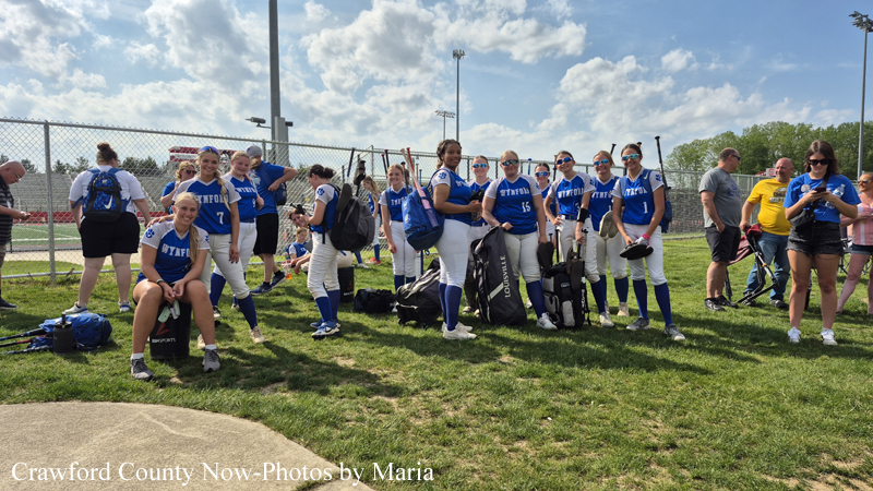 Girls softball team in blue uniforms standing and posing with bags on a grassy field near a chain-link fence.