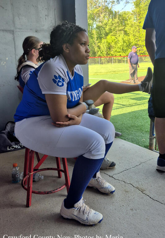 Softball players rest in the dugout; a player in blue and white stretches her leg while seated on a red stool, with teammates nearby.
