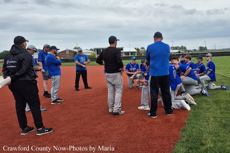 Coach leads a huddle as players in blue jerseys kneel on the red infield during practice on a baseball/softball field.