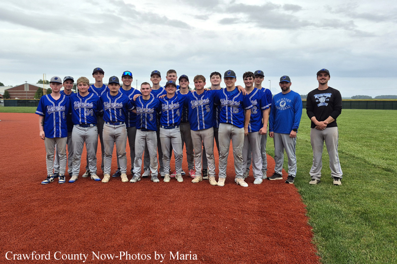 Group of teenage baseball players in blue uniforms posing for a team photo on a baseball field, with coach in darker clothing at the far right.