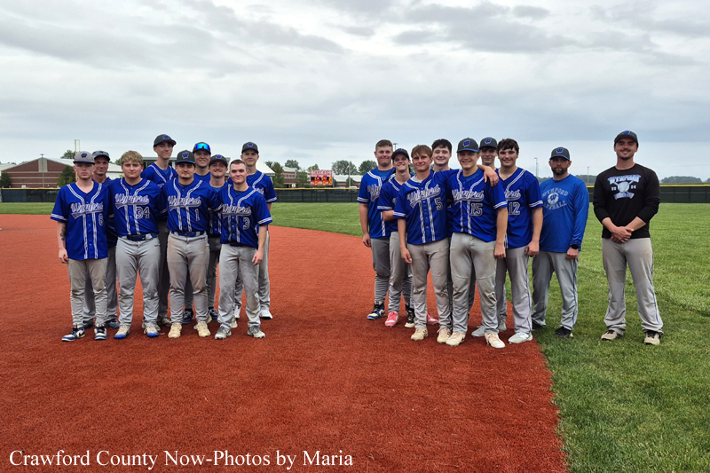Group of young baseball players in blue jerseys posing for a team photo on the infield, with a coach at far right on the grass field behind.
