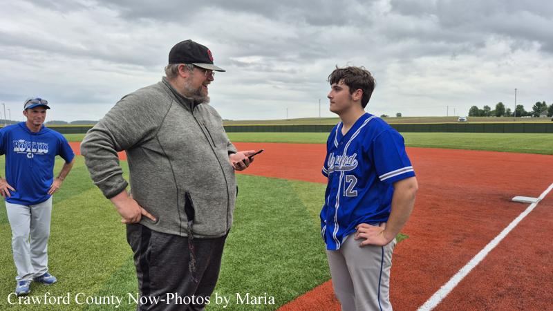 Coach in a gray hoodie talks to a young baseball player in blue jersey on a diamond, cloudy sky overhead