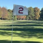 Flag marking hole number 2 on a sunny golf course with green grass and trees in the distance.