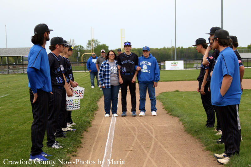 Softball players in blue uniforms form two lines along the dirt infield, facing each other as two people stand in the center posing for a photo on a sunny baseball/softball field.