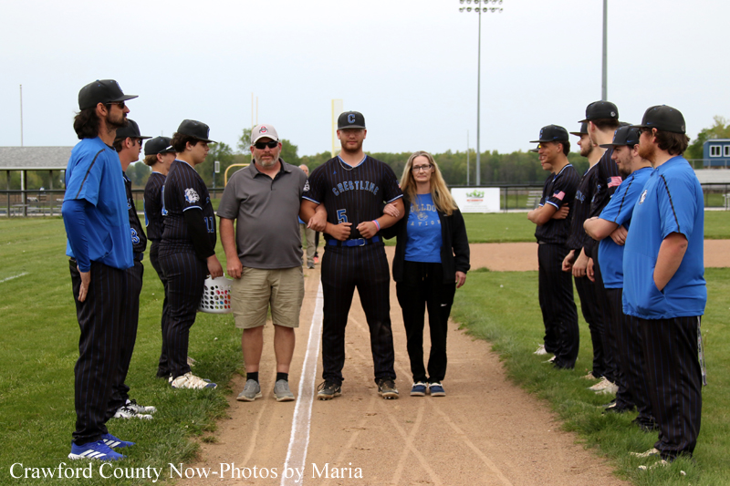 Baseball players form a pre-game lineup along the dirt baseline, with two guests standing center stage between the teams in blue and black uniforms.