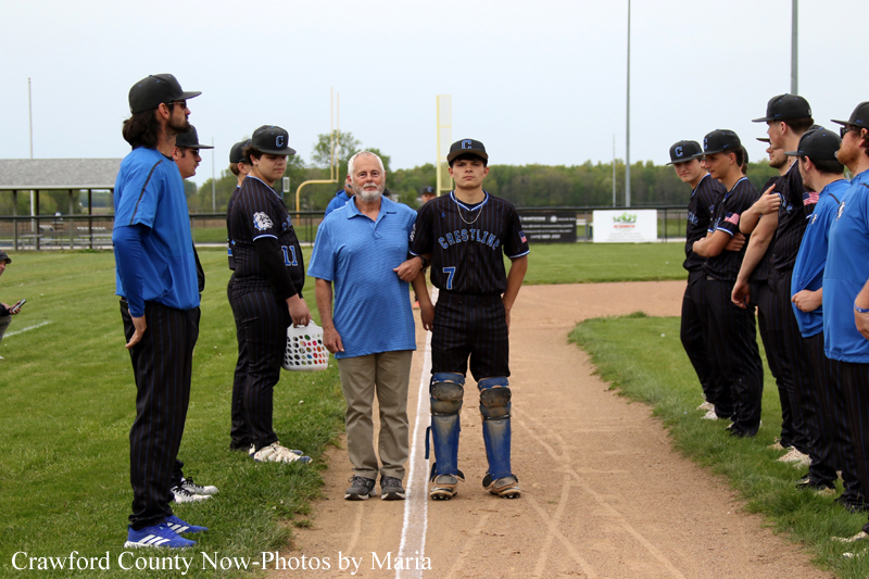Baseball players in black pinstripe uniforms form a lineup on the field, with a catcher and an older man standing center on the dirt baseline.