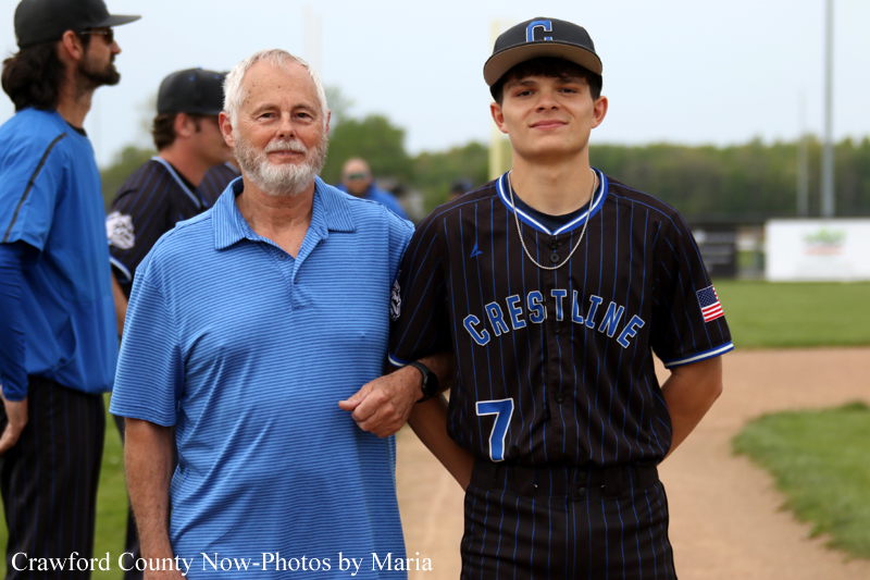 Older man in a blue polo stands with a Crestline baseball player on a dirt baseline at a sunny field, arm linked with the coach's arm around his shoulder.