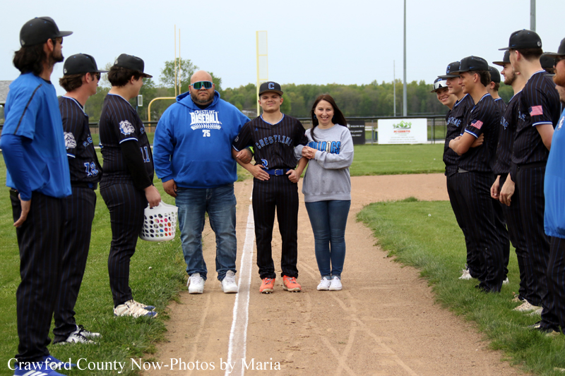 Youth baseball team lines up on a dirt field, with a man in a blue hoodie, a central player in uniform, and a woman in a gray sweatshirt at the center.