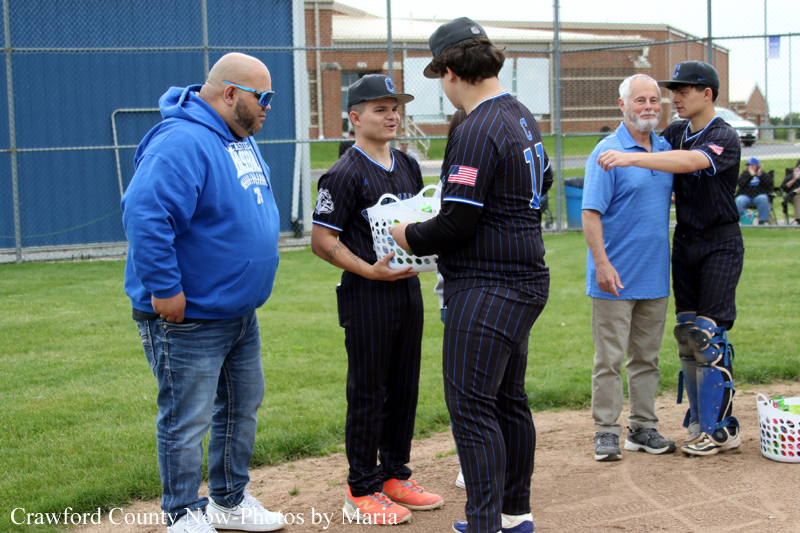 Coach in blue hoodie hands a gear basket to a young baseball player on a grassy field during a game,