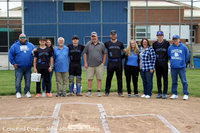 Group of baseball players and coaches posing for a photo on the field in front of a blue building behind them, dusk or daytime event.
