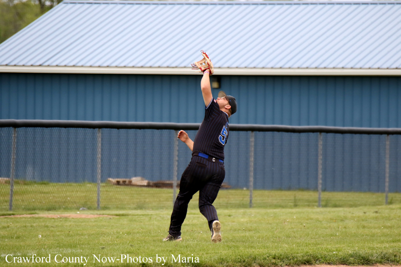 Baseball player in a dark uniform leaps to catch a fly ball with glove raised overhead on a grassy field, blue building in the background