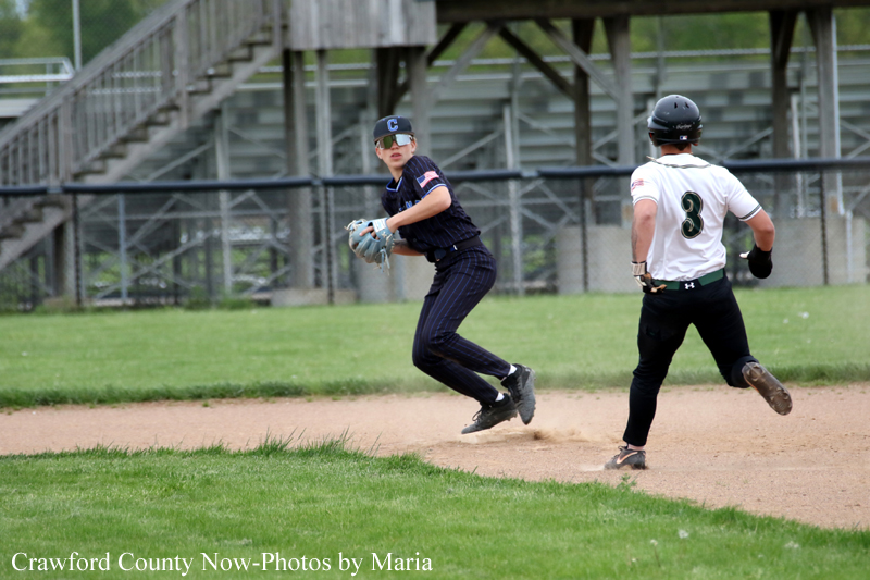 Baseball fielder in a navy uniform with cap watches the play as a baserunner in a white jersey with number 3 runs past the infield.