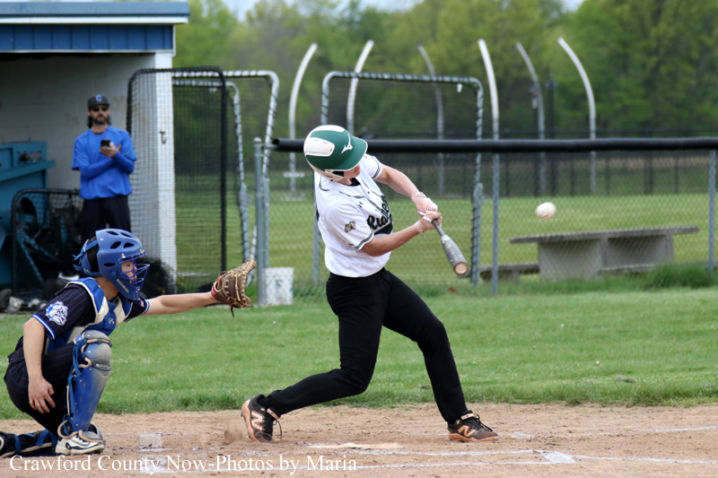 Baseball batter in white and green helmet swings at a pitch, with the catcher in blue gear behind him on the dirt field.