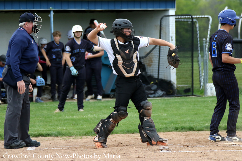 Softball catcher mid-throw in the infield with umpire and teammates watching from the dugout in the background.